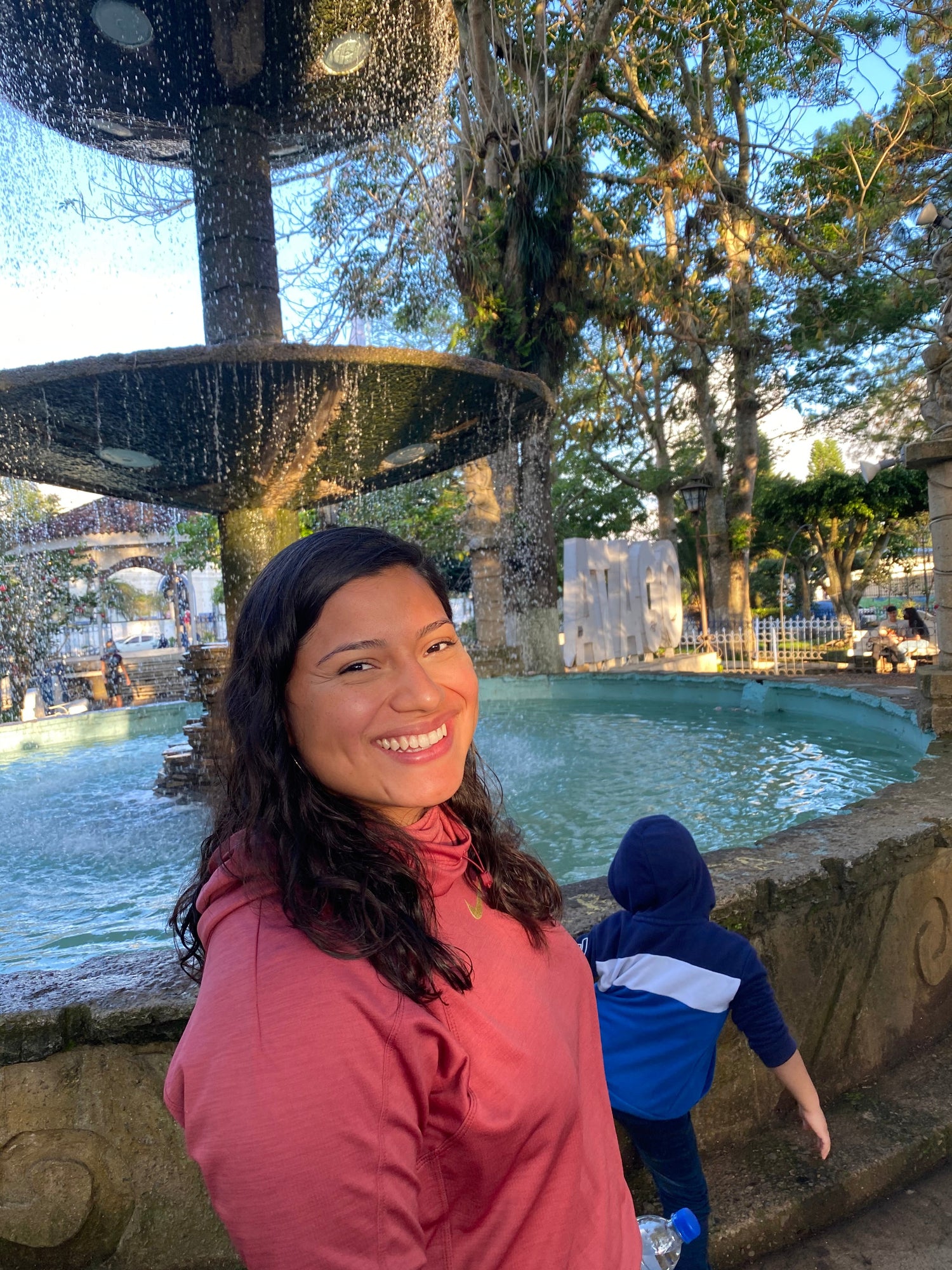 Woman smiling in front of a fountain with trees and people in the background